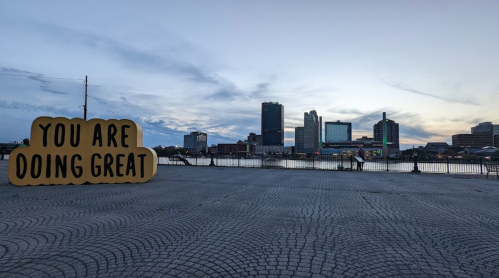 A large sign reading "YOU ARE DOING GREAT" in front of a city skyline at sunset by the water.