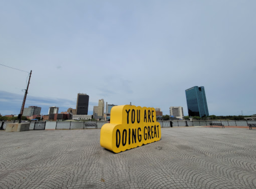 A large yellow sign that reads "YOU ARE DOING GREAT" in an urban setting with a city skyline in the background.