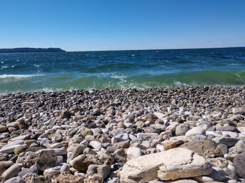 A rocky shoreline with smooth stones, gentle waves, and a clear blue sky in the background.