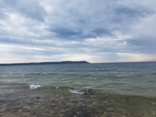 A calm sea under a cloudy sky, with gentle waves lapping against a rocky shore and a distant landform on the horizon.