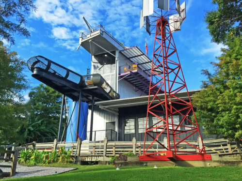 A modern building with a slide, wind turbine, and red tower, surrounded by greenery and a blue sky.