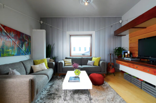 Modern living room with gray sofas, a white coffee table, colorful cushions, and a wooden shelf with a TV.
