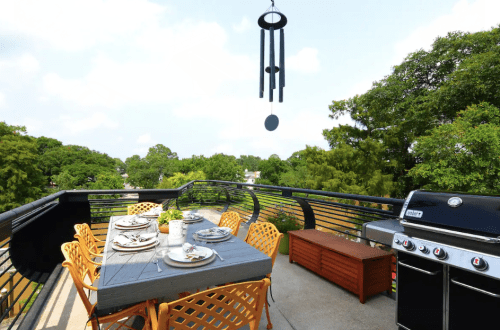 A spacious balcony with a dining table, chairs, a grill, and a wind chime, surrounded by greenery.