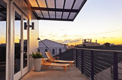 A balcony with two lounge chairs overlooks a sunset, featuring a modern design and city rooftops in the background.