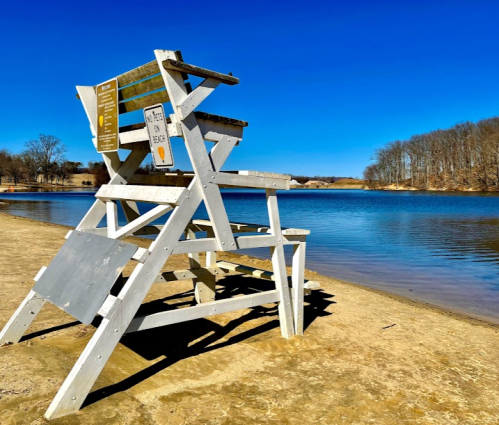 A white lifeguard chair on a sandy beach by a calm lake under a clear blue sky.