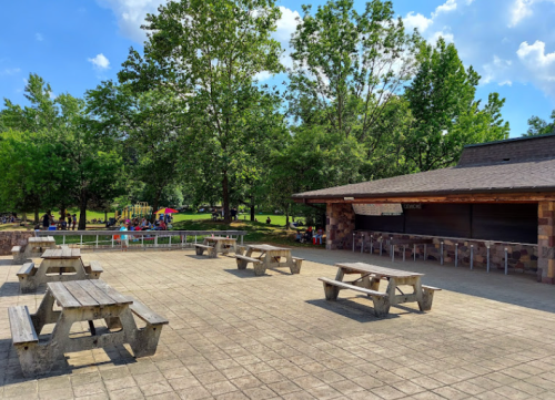 A spacious outdoor area with picnic tables and a concession stand, surrounded by trees and a playground in the background.