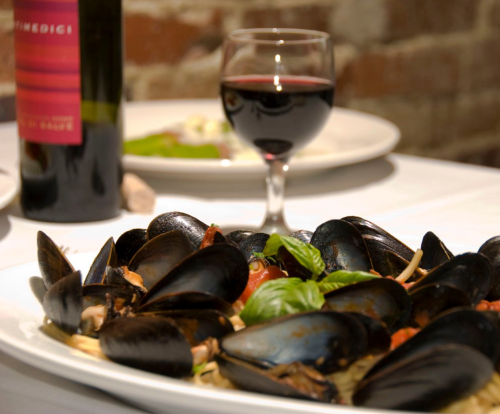 A plate of mussels with pasta, garnished with basil, alongside a glass of red wine and a bottle in the background.