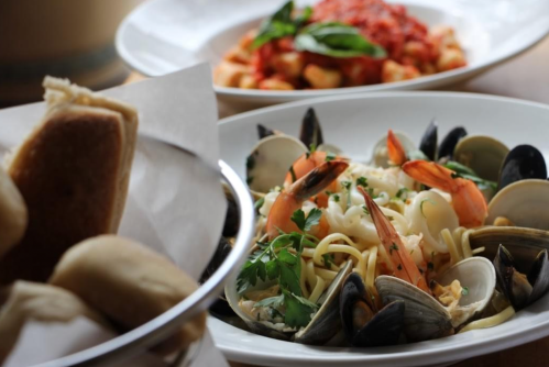 A plate of seafood pasta with shrimp, clams, and herbs, alongside a bowl of gnocchi and bread rolls in the background.