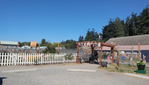 A sunny outdoor area with a wooden entrance, white fence, and buildings in the background, surrounded by trees.