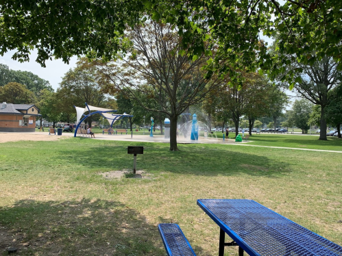 A park scene featuring a blue picnic table, trees, and a splash pad in the background on a sunny day.