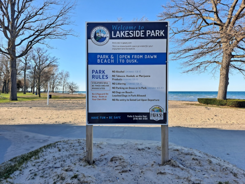 Sign at Lakeside Park detailing park rules and hours, with a view of the beach and lake in the background.