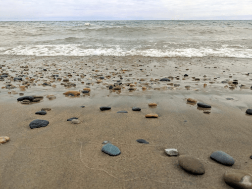 A sandy beach with smooth pebbles in the foreground and gentle waves lapping at the shore under a cloudy sky.