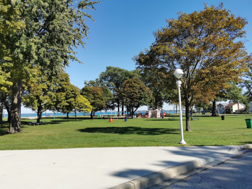 A sunny park with green grass, trees, and a view of the water in the background. A pathway leads through the area.