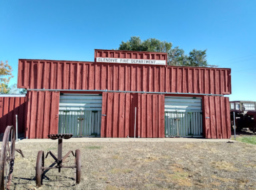 Red wooden building with "Glendive Fire Department" sign, featuring two large garage doors and vintage equipment outside.