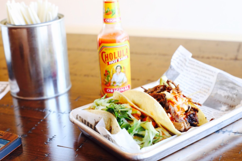 Two tacos filled with meat and toppings, served with a bottle of Cholula hot sauce and a metal container of utensils.