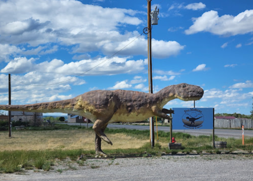 A large dinosaur statue stands next to a sign for a dinosaur-themed attraction under a blue sky with clouds.