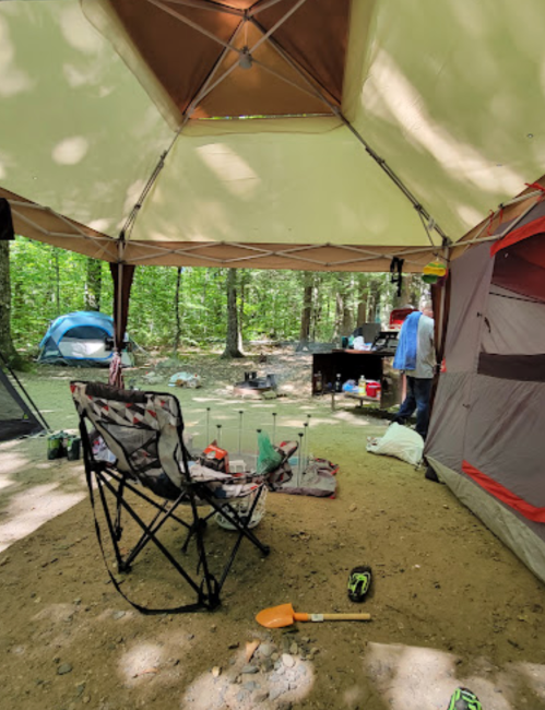 A campsite with a large tent, camping chairs, and a cooking area surrounded by trees and tents in the background.