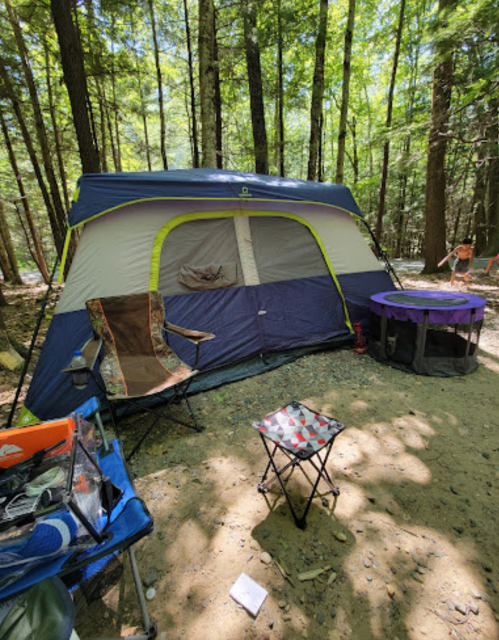A camping setup in a forest, featuring a tent, folding chairs, and a small table on a gravel ground.