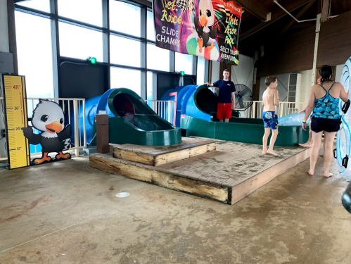 Children playing near water slides in an indoor pool area, with a measuring height chart and colorful decorations.