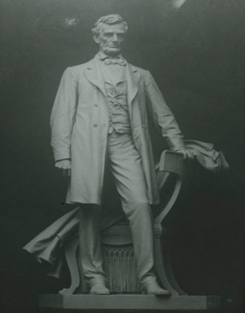 A statue of Abraham Lincoln standing, dressed in a suit with a bow tie, holding a hat, against a dark background.