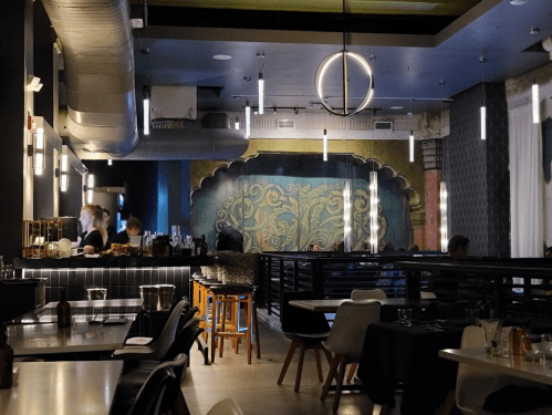 A modern restaurant interior with a bar, artistic wall mural, and stylish lighting, featuring patrons and staff.