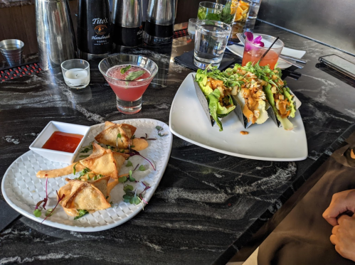 A table with two plates of food: one with samosas and sauce, the other with tacos, alongside drinks.