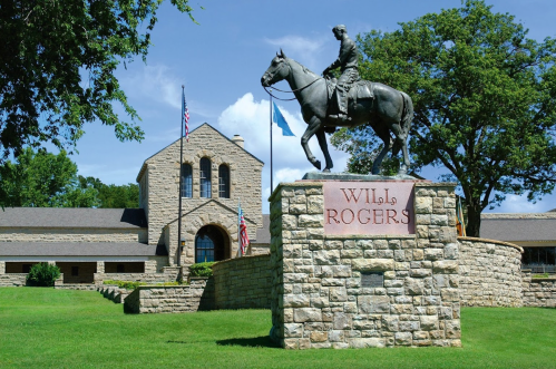 Statue of Will Rogers on horseback in front of a stone building with flags and green lawn.