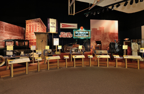 A museum exhibit featuring historical artifacts, including vehicles and signage, set against a vintage backdrop.