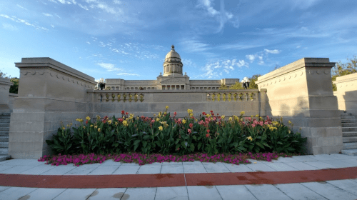 A landscaped area with colorful flowers in front of a grand building under a blue sky with scattered clouds.