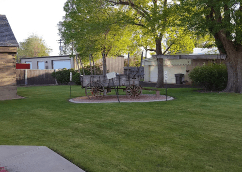 A vintage wooden wagon on display in a grassy area, surrounded by trees and buildings.