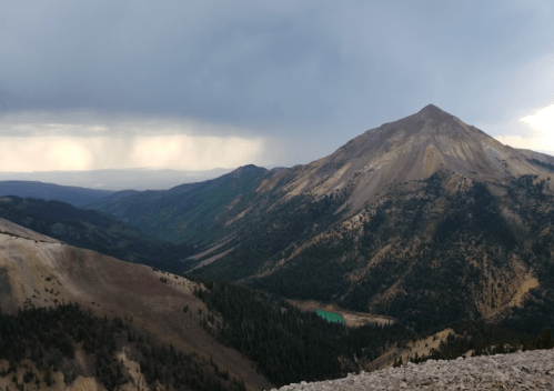 A rugged mountain peak rises above a lush valley under a cloudy sky, with distant hills visible in the background.