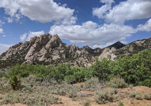 Rocky mountains rise against a blue sky with fluffy clouds, surrounded by green shrubs and dry terrain.
