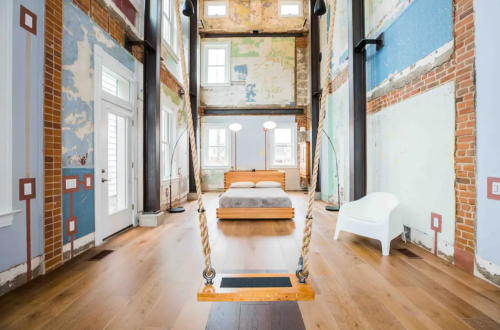 A spacious room with exposed brick walls, a bed, and a swing hanging from the ceiling, featuring large windows.