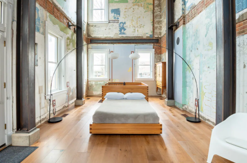 A minimalist bedroom with a wooden bed, large windows, and colorful, peeling wall paint.
