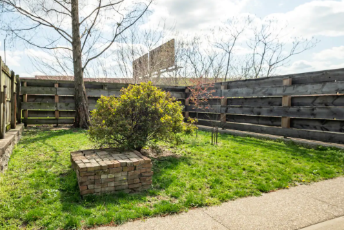 A small grassy area enclosed by wooden fences, featuring a bush and a stone block in the center under a cloudy sky.