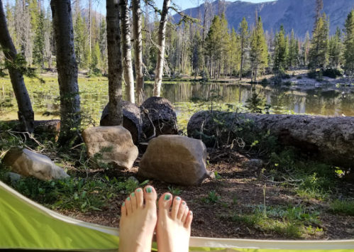 A pair of bare feet with green nail polish resting outside a tent, overlooking a serene lake and forested landscape.