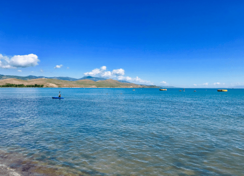 A serene view of a calm blue sea with distant hills and a clear sky, featuring a person paddleboarding.