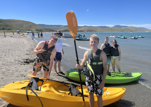 Two young men in life jackets pose on a beach with kayaks, smiling and holding a paddle, with a scenic backdrop.