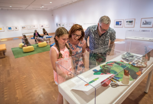 A family examines an art display in a gallery, surrounded by other visitors and framed photographs on the walls.