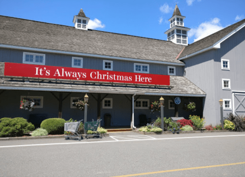 A gray building with a red sign reading "It's Always Christmas Here," featuring flower pots and shopping carts outside.