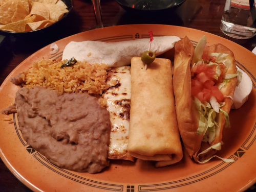 A plate of Mexican food featuring rice, refried beans, a cheese enchilada, a crispy taco, and a soft taco with toppings.