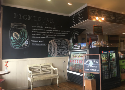 Interior of a cozy café with a chalkboard menu, pickle-themed decor, and a display fridge.