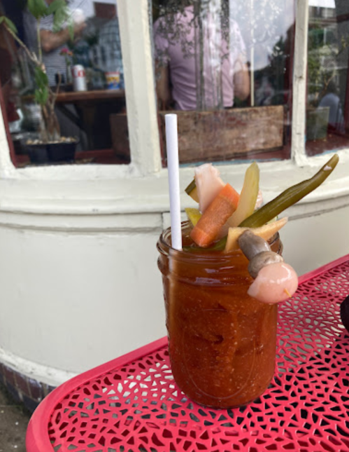 A cocktail in a jar, garnished with pickled vegetables, sits on a red table outside a café.