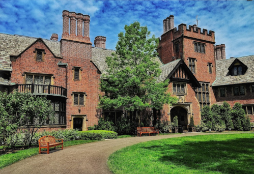 A large brick mansion with a tower, surrounded by greenery and benches, under a blue sky with clouds.