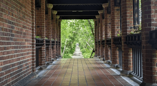 A brick walkway leads through a covered archway, surrounded by lush greenery on both sides.