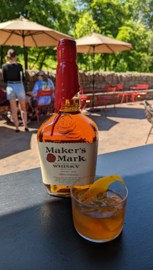 A bottle of Maker's Mark whisky next to a glass of whisky with ice and a lemon twist, set outdoors with tables and umbrellas.