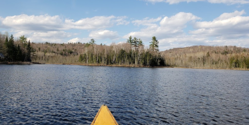 A view from a canoe on a calm lake, surrounded by trees and hills under a partly cloudy sky.