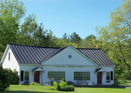 A white building with a black metal roof, featuring large windows and red doors, surrounded by greenery.