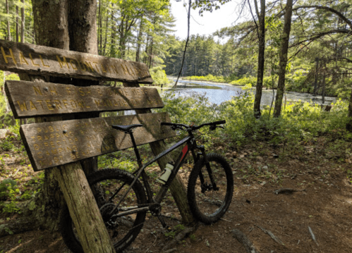 A mountain bike leans against a wooden sign near a serene pond surrounded by trees.