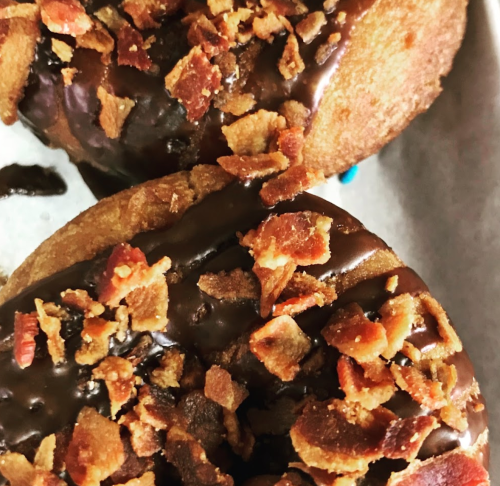 Two donuts topped with chocolate glaze and crispy bacon bits, placed on a white paper background.
